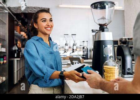 Sorridente proprietario di una caffetteria che tiene il terminale POS e guarda il cliente. Cliente irriconoscibile che paga senza contanti con una carta di credito in un caffè. Foto Stock