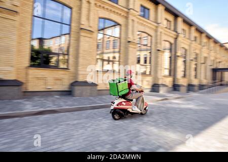 Consegna rapida durante l'epidemia di covid 19. Vista posteriore di un corriere maschile con zaino termico indossando una maschera protettiva nera guidando scooter lungo la strada, fuoco selettivo su un uomo. Concetto di soggiorno a casa Foto Stock