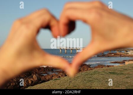 Molo di Port Elizabeth Shark Rock chiuso da fuori fuoco mani che fanno la forma di un cuore Foto Stock