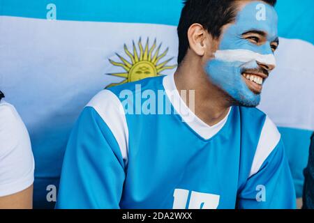 Uomo seduto nello stadio con il viso dipinto in colori argentini. Devoto fan con bandiera argentina dietro nello stadio di calcio. Foto Stock
