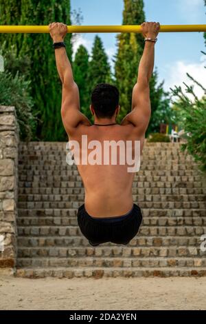 Primo piano ritratto da dietro il giovane latino senza camicia e pantaloni neri e maschera di allenamento al bar di un parco calistenico Foto Stock