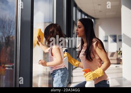 Donna con la figlia che pulisce i vetri usando lo spray insieme Foto Stock