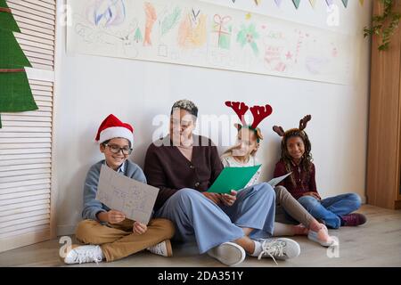 Ritratto a lunghezza intera di insegnante femminile seduto sul pavimento con gruppo multi-etnico di bambini che tengono le immagini mentre godendo la lezione d'arte su Natale, copy spazio Foto Stock