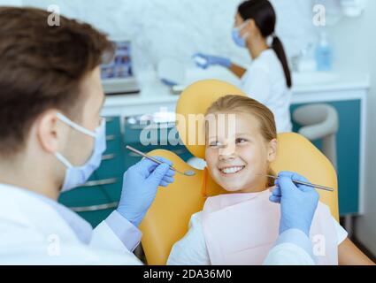 Medico maschile in camice bianco con guanti di gomma e protettivo la maschera tratta i denti della ragazza sorridente in sedia Foto Stock