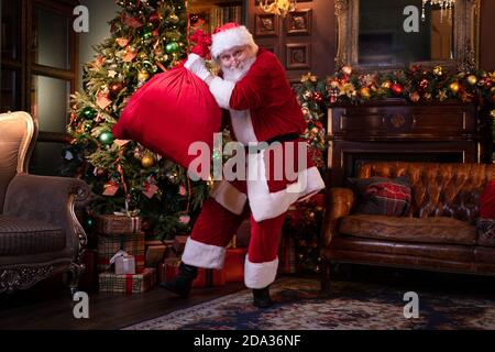 Babbo Natale che si prepara a congratularsi con i bambini. Felice Babbo Natale ascoltando musica e ballando con borsa di regali a casa vicino all'albero di Natale Foto Stock