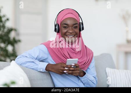 Allegro nero musulmano femmina ascoltare musica sullo smartphone a casa, indossando cuffie Foto Stock