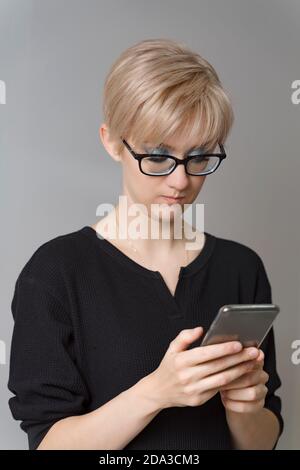 bella giovane donna bionda in occhiali guarda emotivamente allo smartphone. Foto Stock