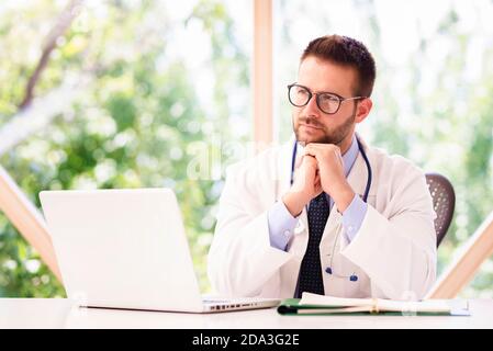 Un medico di pensiero seduto dietro il computer portatile mentre lavora in ufficio medico. Foto Stock