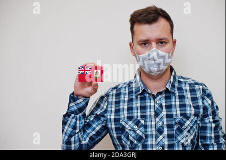 Uomo in camicia a scacchi mostra carta bandiera Bermuda in mano, indossare maschera di protezione isolato su sfondo bianco. I paesi americani concetto di Coronavirus. Foto Stock