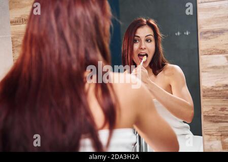 Bella giovane donna in piedi in bagno vicino allo specchio e. spazzolando i denti Foto Stock