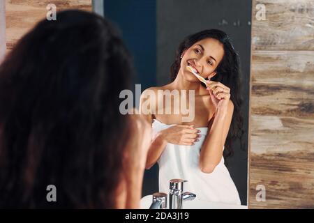 Bella giovane brunetta in piedi in bagno vicino allo specchio e. spazzolando i denti Foto Stock