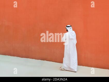 Uomo arabo con abiti tradizionali emirates che camminano all'aperto nel strada contro un muro arancione sfondo - etnia araba e. religione musulmana conc Foto Stock