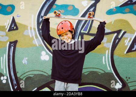 Ritratto di un giovane skater che indossa un casco rosso e gioca con un vecchio skateboard, di fronte a un murale di graffiti Foto Stock