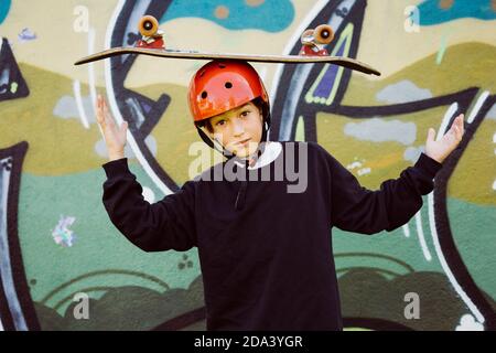 Ritratto di un giovane skater che indossa un casco rosso e gioca con un vecchio skateboard, di fronte a un murale di graffiti Foto Stock