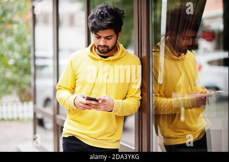 Giovane hipster cittadino indiano in una felpa gialla alla moda. Cool sud asiatico ragazzo indossare felpa con cappuccio guardato telefono cellulare. Foto Stock