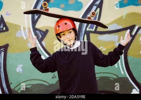 Ritratto di un giovane skater che indossa un casco rosso e gioca con un vecchio skateboard, di fronte a un murale di graffiti Foto Stock