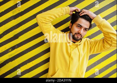 Giovane hipster cittadino indiano in una felpa gialla alla moda. Cool ragazzo del sud asiatico indossare felpa con cappuccio contro lo sfondo a righe. Foto Stock