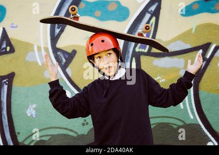 Ritratto di un giovane skater che indossa un casco rosso e gioca con un vecchio skateboard, di fronte a un murale di graffiti Foto Stock