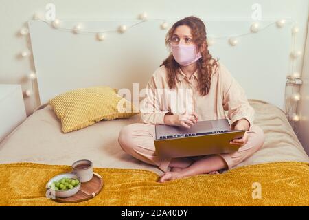 Una donna in una maschera medica lavora online con un computer portatile su un letto di casa. Lavoro remoto da casa durante il blocco a causa del coronavirus. Foto Stock