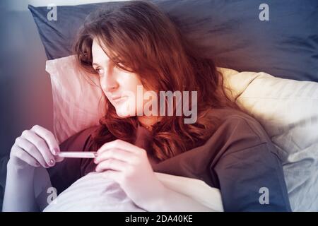 Donna rossa con virus influenzale con maschera sul viso guarda un termometro. Una donna con infezione da coronavirus si trova a letto Foto Stock