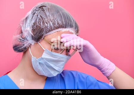 Donna medico in una maschera medica su uno sfondo rosa tiene stiramente su una testa irritata. Medica in disperazione, concetto di epidemia di coronavirus. Foto Stock