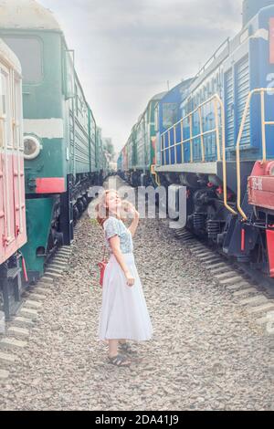 I treni sono sui binari. Giovane donna tra due locomotive. Il fumo proveniente dal motore del treno. Ragazza vicino alle rotaie in tempo nuvoloso. Dangerou Foto Stock