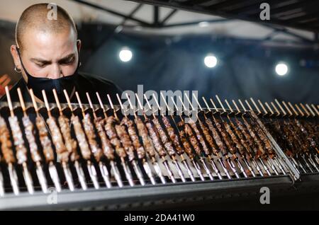 Cuneo, Italia - 3 ottobre 2020: Arrosticini tradizionali abruzzesi (spiedini tipicali) cucinati di notte da un giovane che indossa una maschera protettiva su una stre Foto Stock