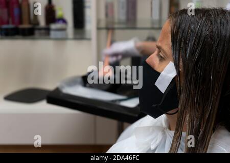 Donna cliente indossare maschera protettiva contro il coronavirus in parrucchiere, parrucchiere tintura capelli cliente con un pennello..New stile di vita normale conc Foto Stock