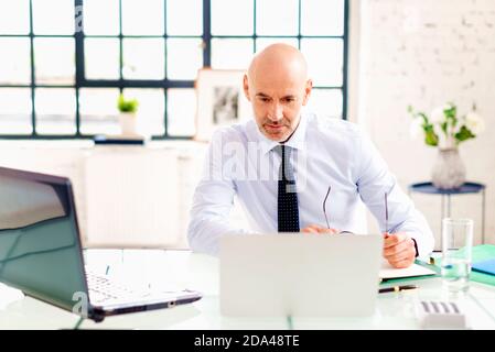 Ritratto di un uomo d'affari anziano infelice seduto alla scrivania e che utilizza un computer portatile mentre lavora in ufficio. Foto Stock
