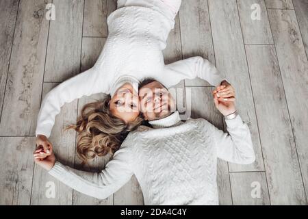 Coppia sorridente in pullover sul pavimento. Felice coppia amorevole in maglia bianca maglie invernali giacenti sul pavimento tenendo le mani. Foto Stock