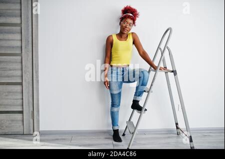 Donna afroamericana in singlet giallo e occhiali da vista contro parete bianca stand om scala in ferro. Foto Stock