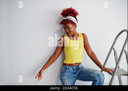 Donna afroamericana in singlet giallo e occhiali da vista contro parete bianca stand om scala in ferro. Foto Stock