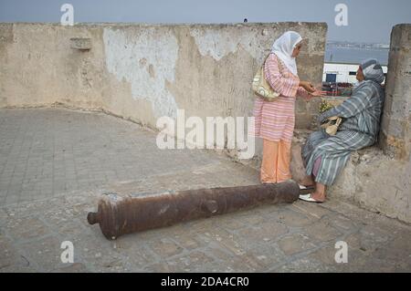 El Jadida, Marocco 2007 agosto. Due donne chiacchierano lungo le mura della città accanto a un vecchio cannone di ferro. Foto Stock