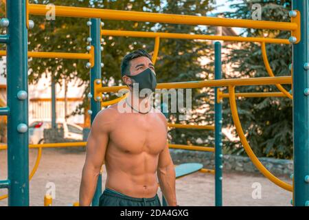 Giovane uomo latino in pantaloni neri e maschera facendo esercizi in un parco calistenico senza t-shirt Foto Stock