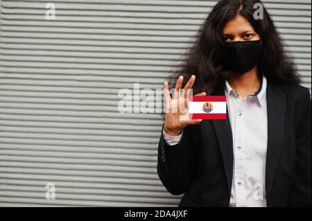 Donna asiatica con abbigliamento formale e maschera protettiva nera tenere la bandiera della Polinesia francese a portata di mano su sfondo grigio. Coronavirus al concetto di paese. Foto Stock