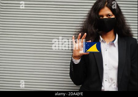 Donna asiatica con abbigliamento formale e maschera protettiva nera tenere la bandiera Tokelau a portata di mano su sfondo grigio. Coronavirus al concetto di paese. Foto Stock