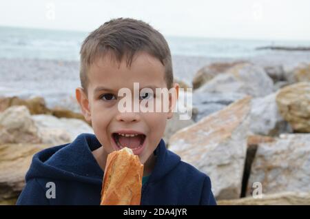 Il ragazzo mangia baguette in tuta blu scuro. Ragazzo sta dipingendo mentre si siede su una spiaggia di pietra sulla riva del mare. Foto Stock