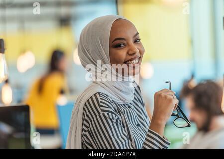 Protarit di una donna musulmana nera (afro-americana) in piedi in un moderno ufficio d'affari mentre indossa un hijab. Foto Stock