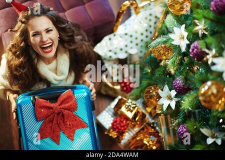 Buon Natale. Vista superiore di felice trendy 40 anni donna in gonna di paillettes oro e maglione con borsa trolley e grande arco rosso vicino albero di Natale A. Foto Stock