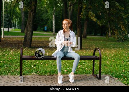 Giovane donna sportiva dai capelli rossi che indossa abiti sportivi seduti sulla panca e tenere gli auricolari nelle mani Foto Stock