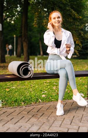 Ritratto di giovane donna allegra e sportiva seduta sulla panca nel parco cittadino e indossando le cuffie. Foto Stock