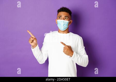 Concetto di coronavirus, quarantena e stile di vita. Uomo afro-americano scioccato, indossando maschera medica, puntando e guardando nervoso in alto a sinistra Foto Stock