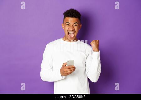 Immagine di felice afroamericano uomo, vincendo qualcosa in linea, tenendo il telefono cellulare e trionfando, dicendo sì con gioia, in piedi su viola Foto Stock