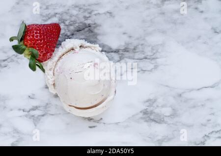 Vista su un piatto di gelato alla fragola rosa con una sola frutta fresca alla fragola su uno sfondo di marmo bianco e grigio. Vista dall'alto. Foto Stock