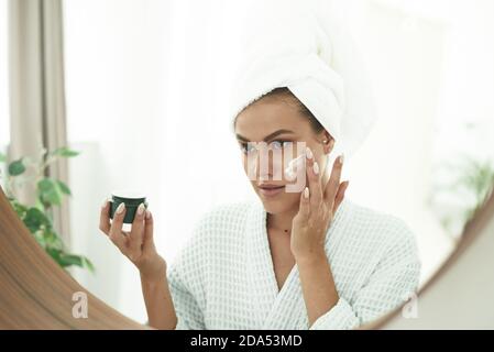 Una bella giovane donna con vitiligine sulle sue mani in un accappatoio e con un asciugamano sulla sua testa applica moisturizer alla sua faccia. Il concetto di cura della pelle, idratazione e pulizia del viso e del collo. Foto Stock