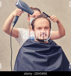 Un parrucchiere in una maschera medica con un asciugacapelli fa styling dei capelli ad un cliente in una stanza da bagno domestica Foto Stock