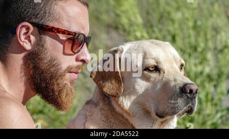 Biondo uomo ispanico con occhiali da sole che guarda all'orizzonte con il suo labrador retriever Foto Stock