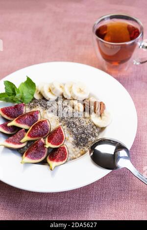 una ciotola con porridge di farina d'avena con semi di chia superfood, fichi freschi e banana su sfondo rosa, servita con tè nero Foto Stock