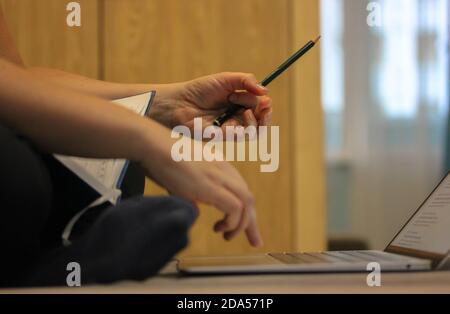 Una donna lavora a distanza da casa, digitando sul suo computer, tenendo una matita verde in mano e un diario aperto in grembo. Foto Stock