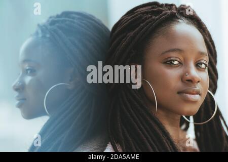 Un ritratto ravvicinato di una giovane donna nera carina con dreadlock e orecchini enormi, piercing nel suo naso, è appoggiata contro una parete di vetro all'aperto Foto Stock